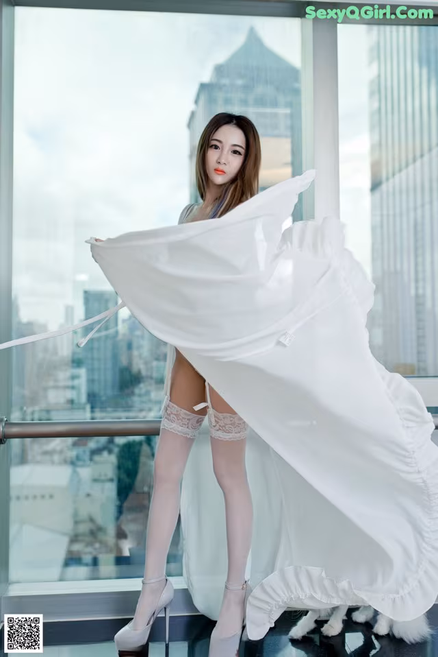 A woman in a white dress and stockings posing by a window.